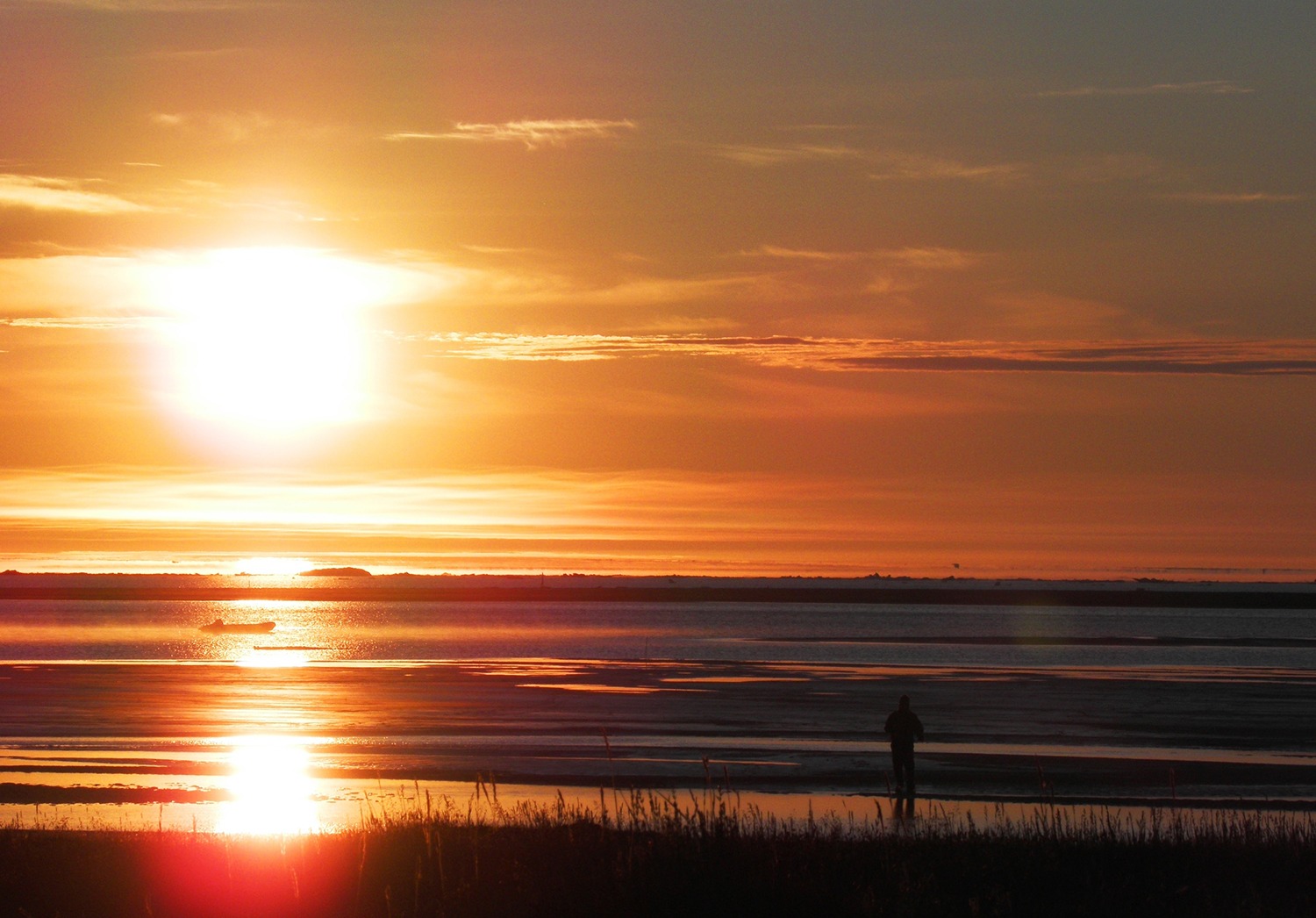 Orange sunset over water and grass with person standing on beach with grass in the foreground.