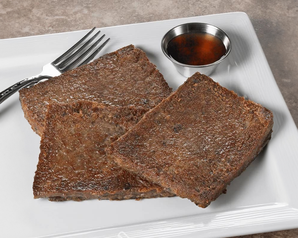 Three slices of crispy fried scrapple on a white plate, accompanied by a small bowl of syrup and a fork.