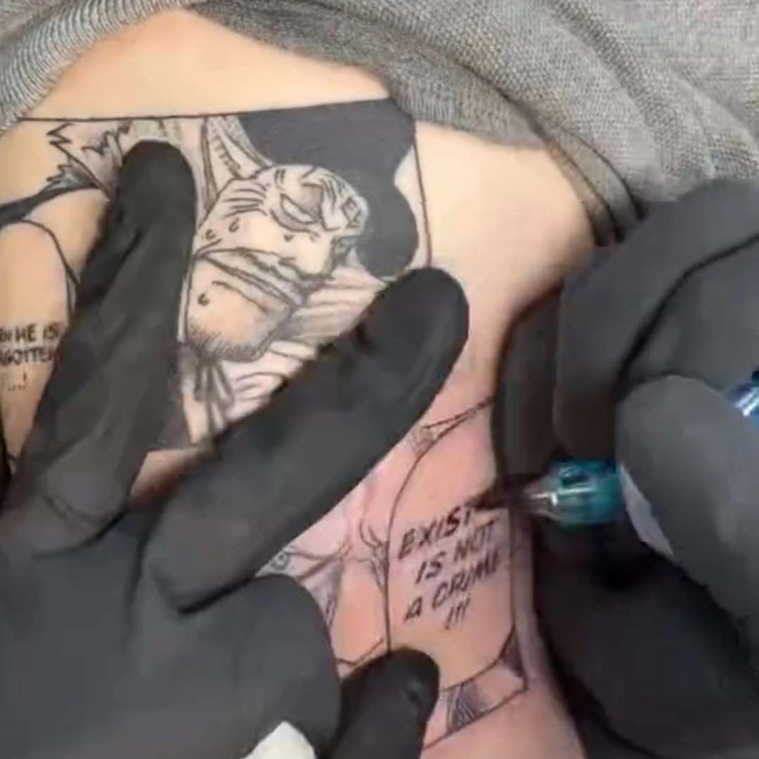 A close-up of a person getting a tattoo. A gloved hand uses a tool to ink an anime-style character and the words "EXIST IS NOT A CRIME" onto skin.