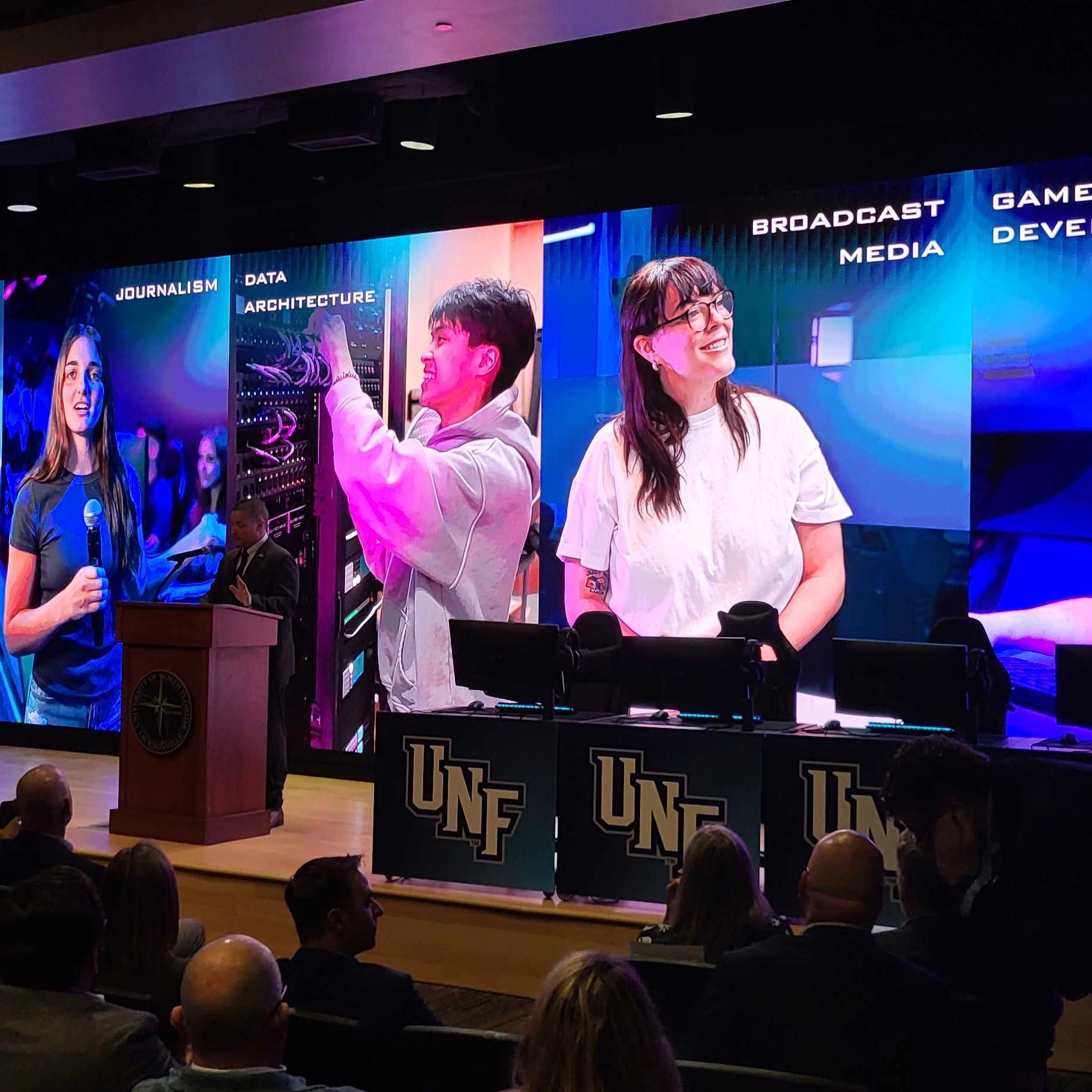 An audience watches a presentation at the UNF Flight Deck. A large screen displays student photos under headings for "JOURNALISM," "DATA ARCHITECTURE," and "BROADCAST MEDIA." A speaker stands at a podium next to stage monitors and desks branded with the UNF logo.
