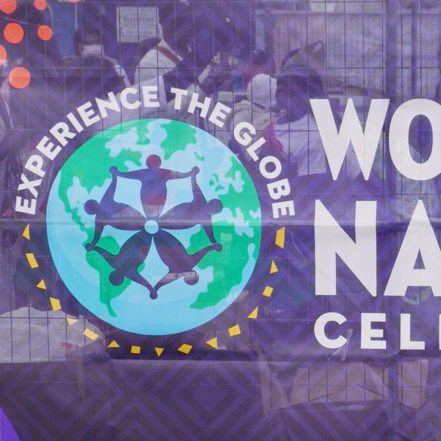 A banner for the World of Nations festival features a circular logo with a globe and figures holding hands, surrounded by the text "EXPERIENCE THE GLOBE." The word "CELEBRATION" is partially visible on the right. The banner has a purple geometric pattern and is displayed in front of a wire fence.