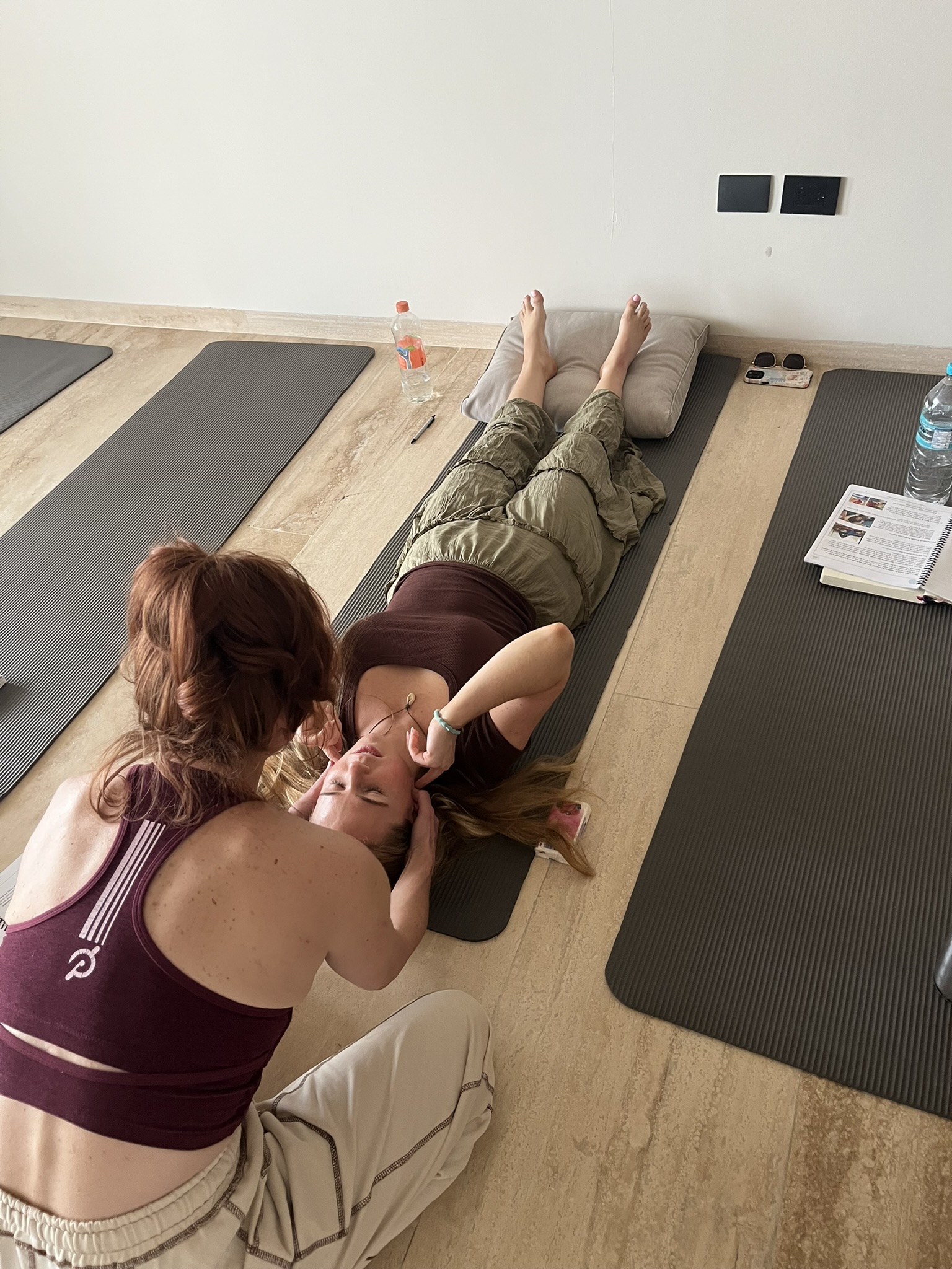 Top-down view of two women on gray yoga mats in a bright studio: one lying on her back with legs raised on a cushion while the other gently supports her head, suggesting a neck or yoga stretch. A water bottle, notebook, pen, sunglasses, and another bottle sit nearby on the floor near the mats.