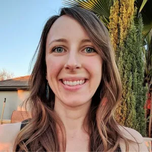 Portrait of a smiling woman with long brown hair outdoors, with greenery and a clear blue sky in the background.