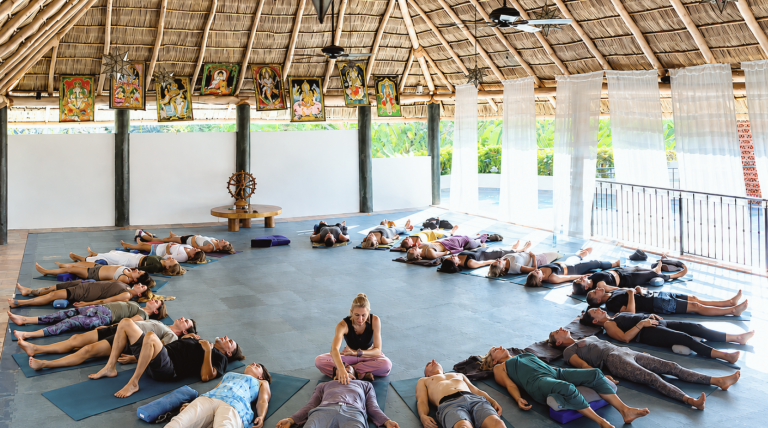 Group of people lying on yoga mats in a circular arrangement inside a thatched-roof, open-air studio; instructor sits cross-legged at center guiding practice.