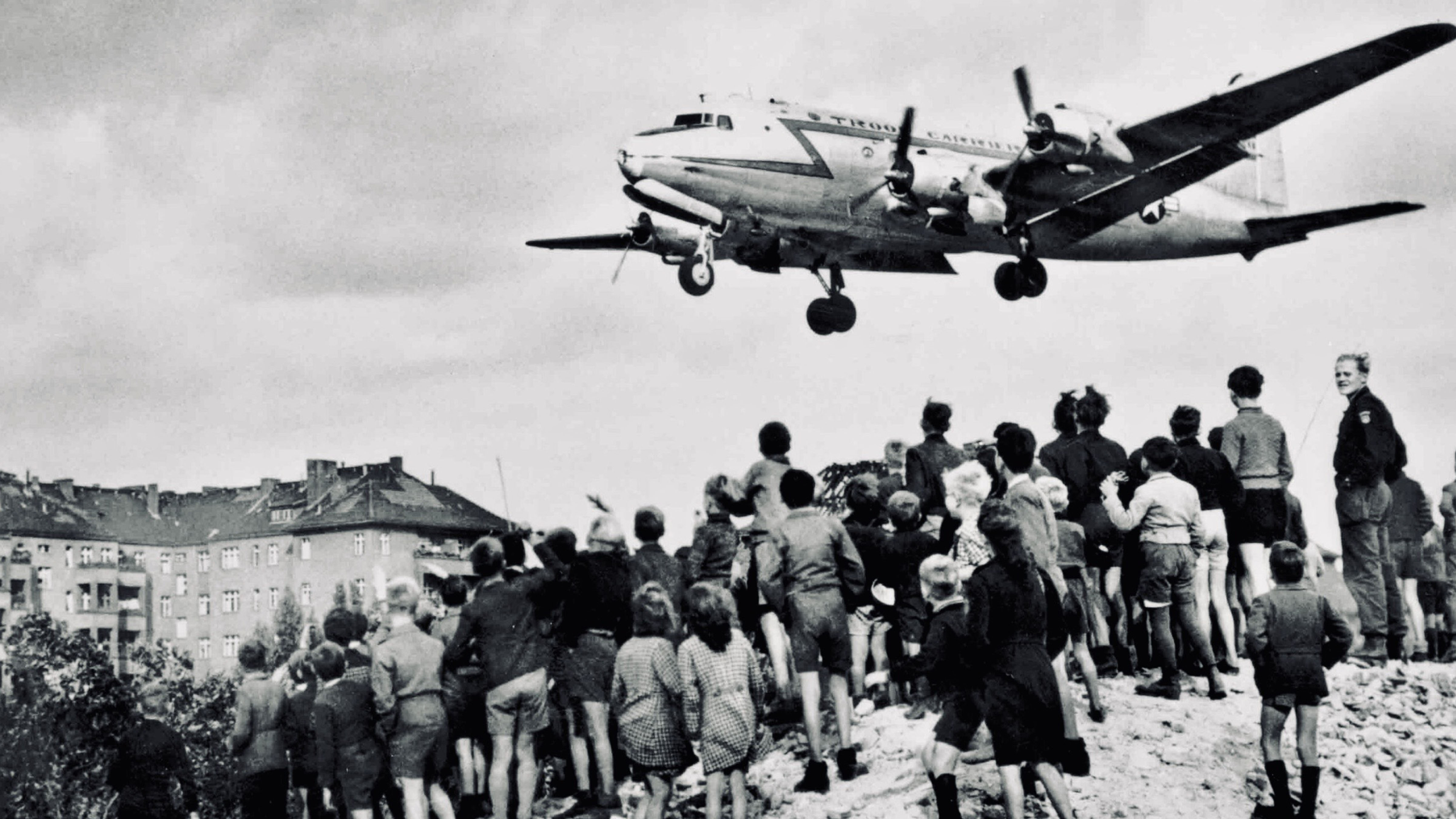 Children stand on a pile of rubble in West Berlin as an Allied transport aircraft approaches during the Berlin Airlift in 1948.