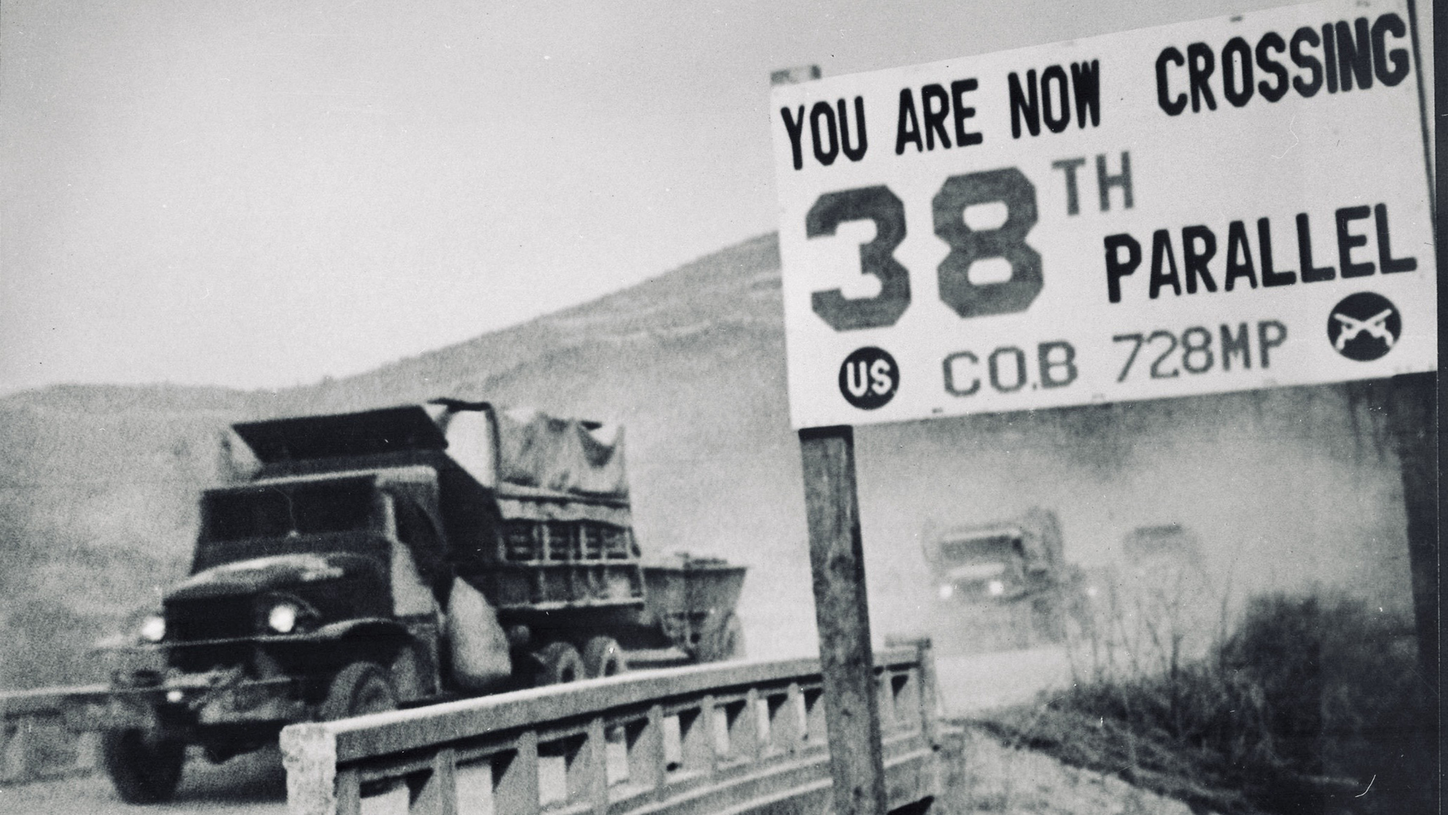 Historic black-and-white photograph of United Nations military trucks crossing the 38th Parallel during the Korean War, passing a roadside sign marking the boundary.