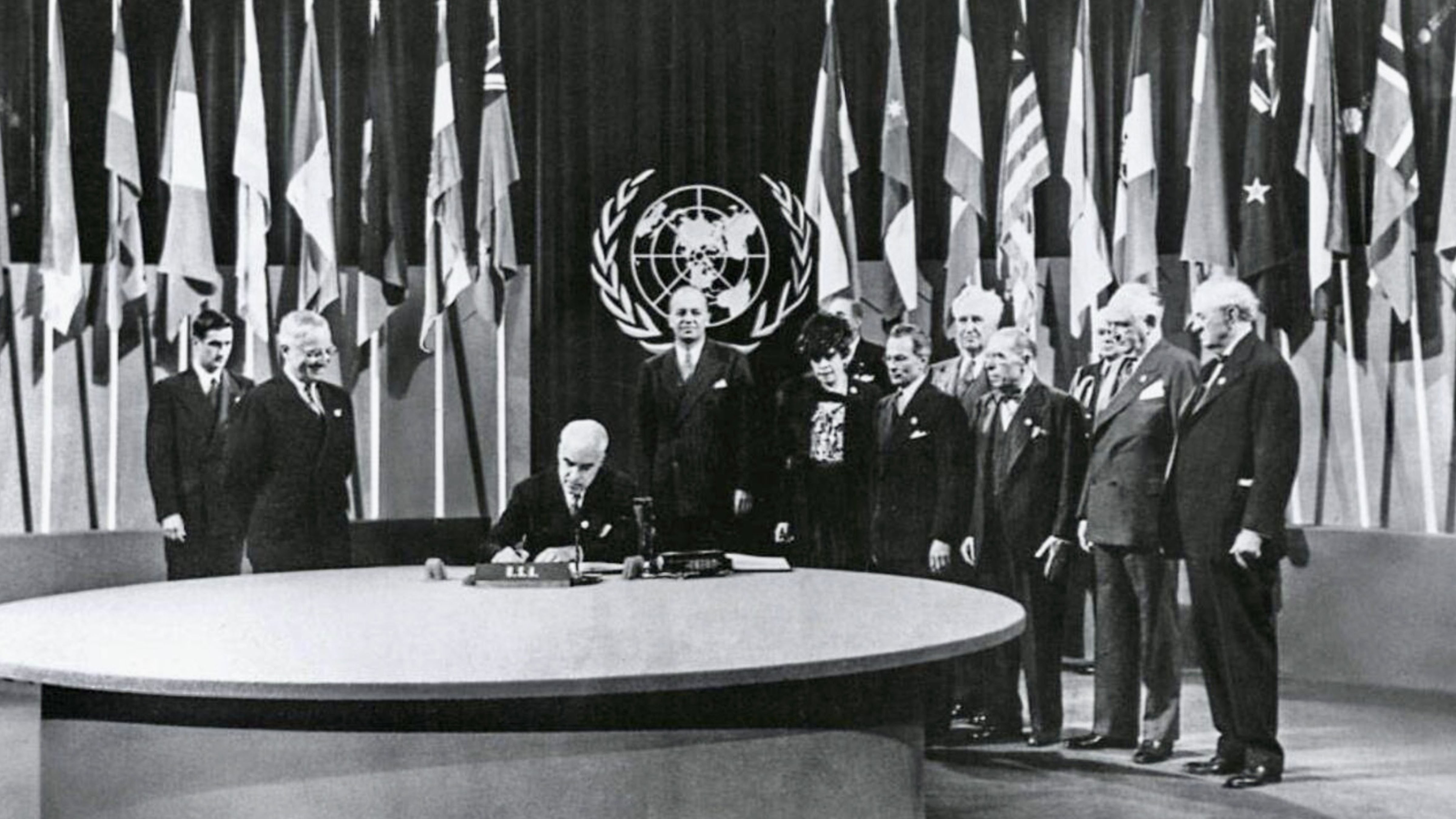 Representatives of a founding member nation sign the United Nations Charter during the San Francisco Conference on June 26, 1945, as delegates from other countries look on beneath the flags of participating nations.