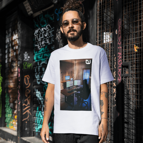 A man with a beard and a top bun, wearing round sunglasses and a white Over-sized t-shirt featuring an "Upcoming Engineer" logo. The t-shirt has a printed image of a person sitting at a computer desk with multiple monitors. The man is standing in an urban environment with graffiti-covered walls, giving the scene a streetwear vibe. The "Upcoming Engineer" logo is placed at the top right corner of the printed image on the t-shirt.