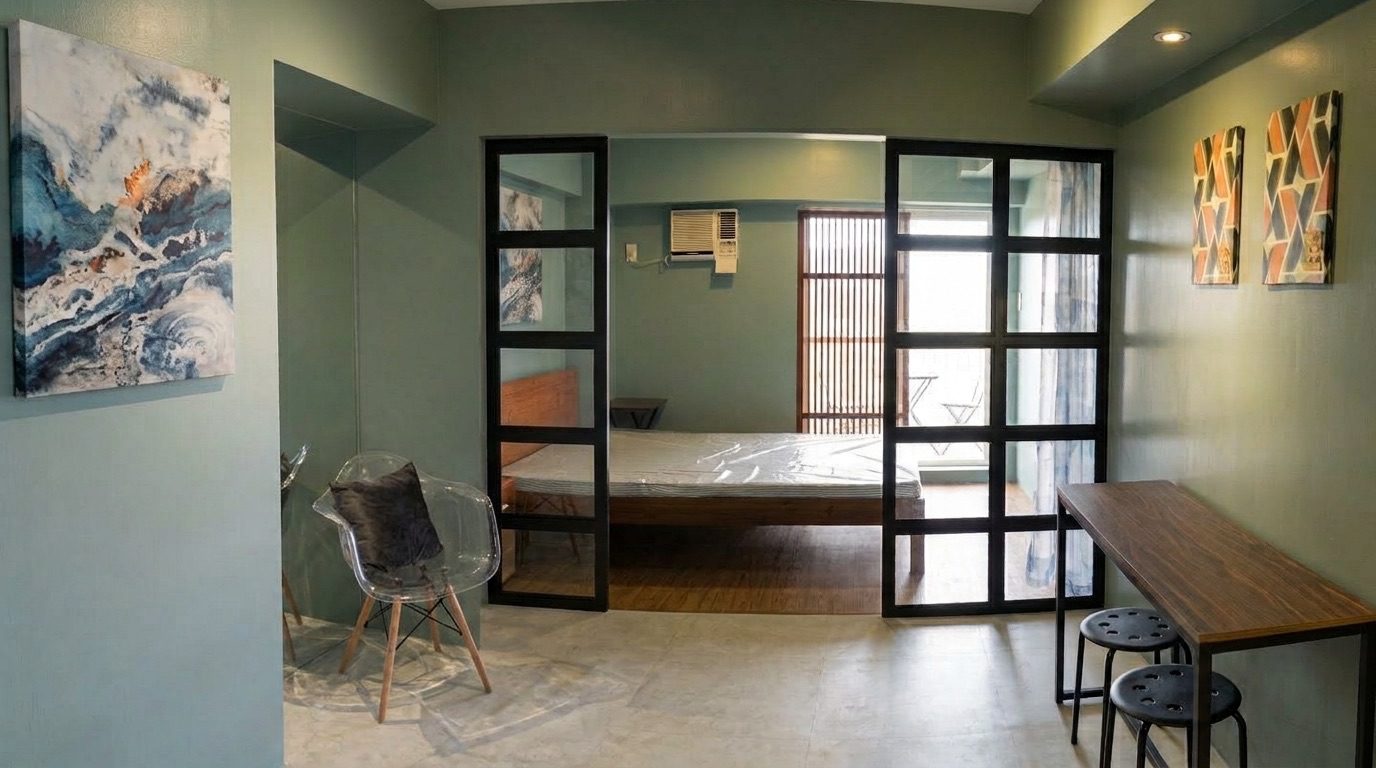 Wide view of the sage-green studio showing the entry area, marble-pattern wall art, dining nook with two stools, and glass sliding doors opening to the bedroom, wood bed frame, and balcony with an unobstructed outdoor view.