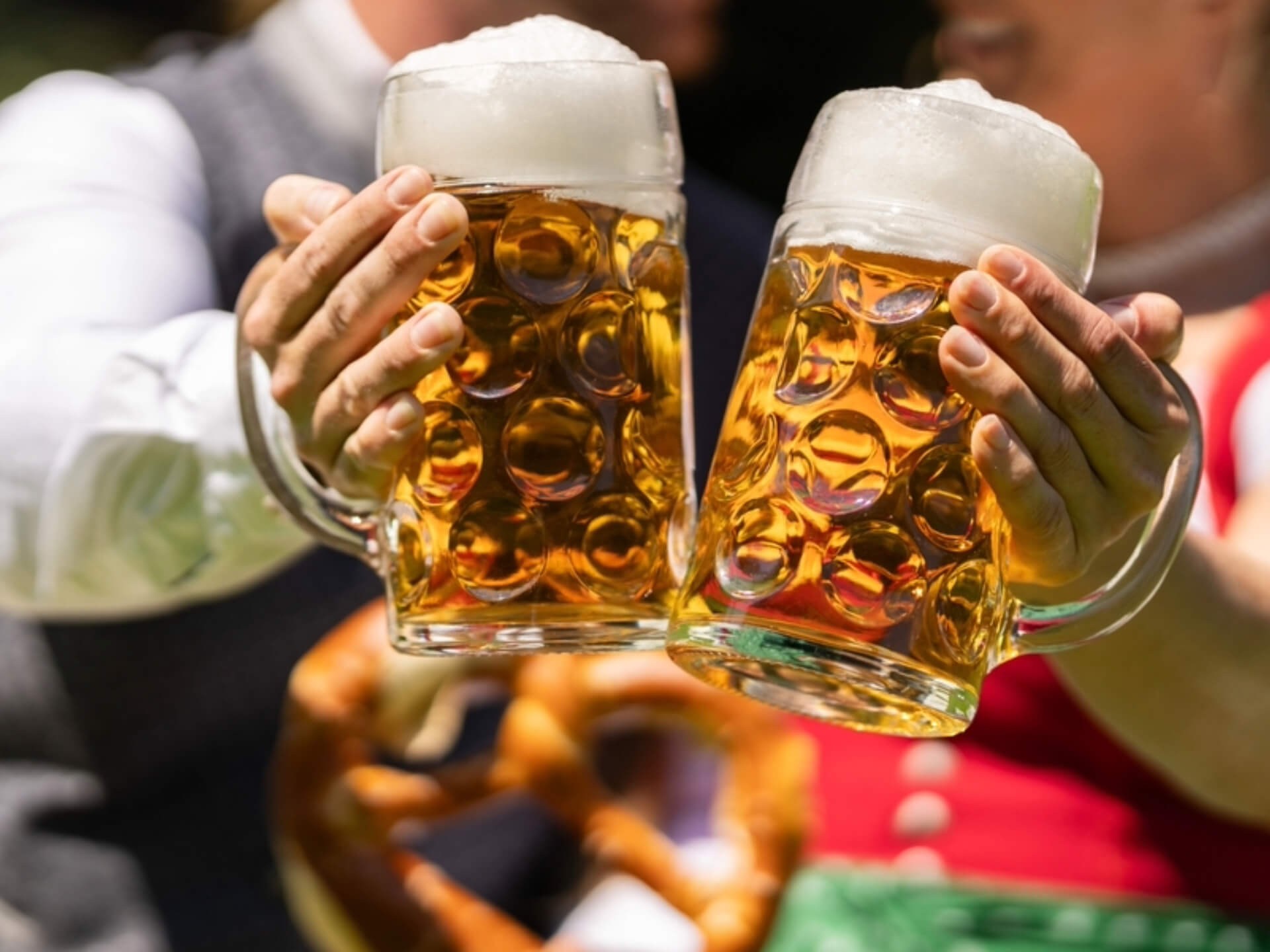 Ein Mann und eine Frau in traditioneller bayerischer Tracht stoßen mit großen, schäumenden Bierkrügen an; im Hintergrund ist eine Brezel zu sehen – auf dem Oktoberfest in Florida