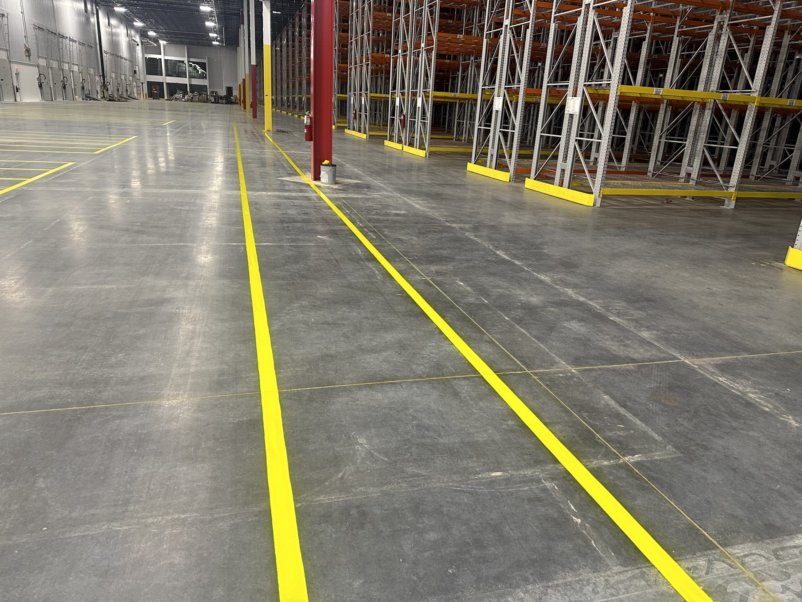 Empty warehouse interior with yellow floor markings, tall storage racks in the background, and a high ceiling with lights.