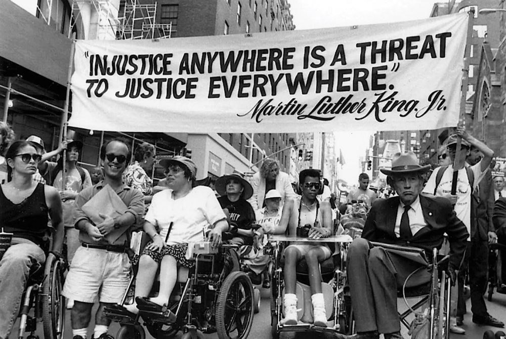 Image: black and white photograph of individuals in wheelchairs, and with other visible disabilities, taking part in a political march. Participants carry a banner that reads: "Injustice Anywhere Is a Threat To Justice Everywhere ~ Martin Luther King Jr."
