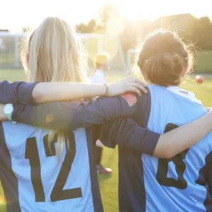 Three soccer players in numbered light blue team jerseys standing arm in arm on a field at sunset