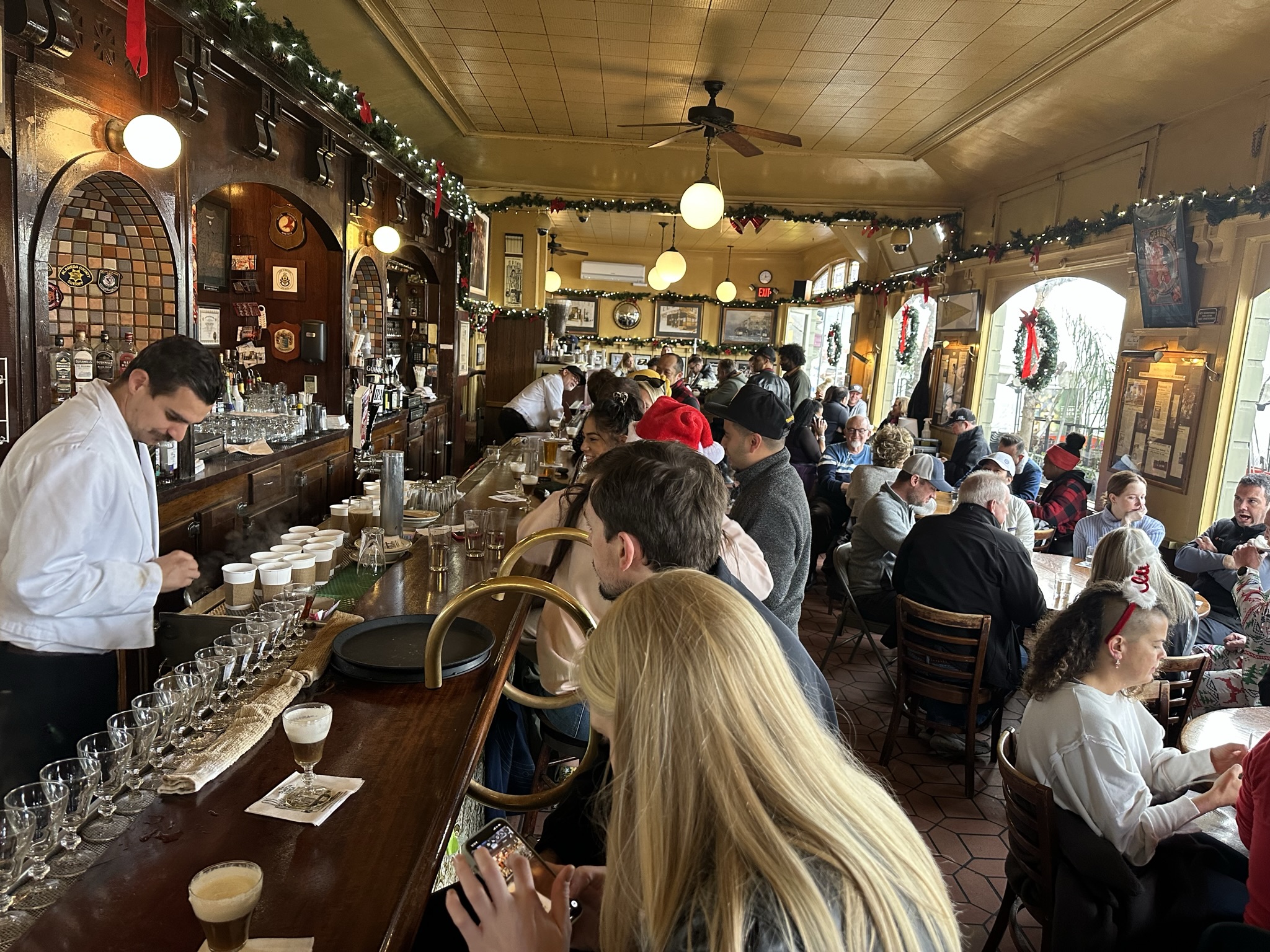 A white-jacketed bartender prepares a row of Irish coffees at a packed historic San Francisco bar during a busy holiday service