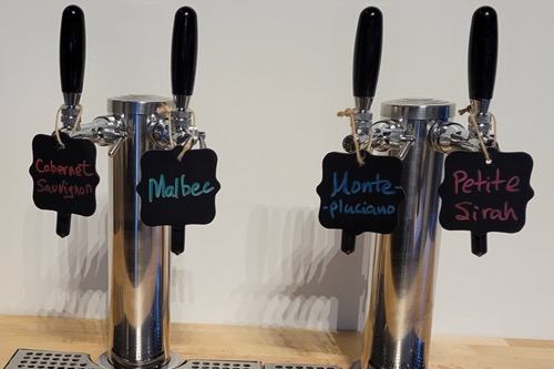 Four stainless wine taps on a tasting room bar, each with a handwritten chalk tag reading Cabernet Sauvignon, Malbec, Montepulciano, and Petite Sirah.