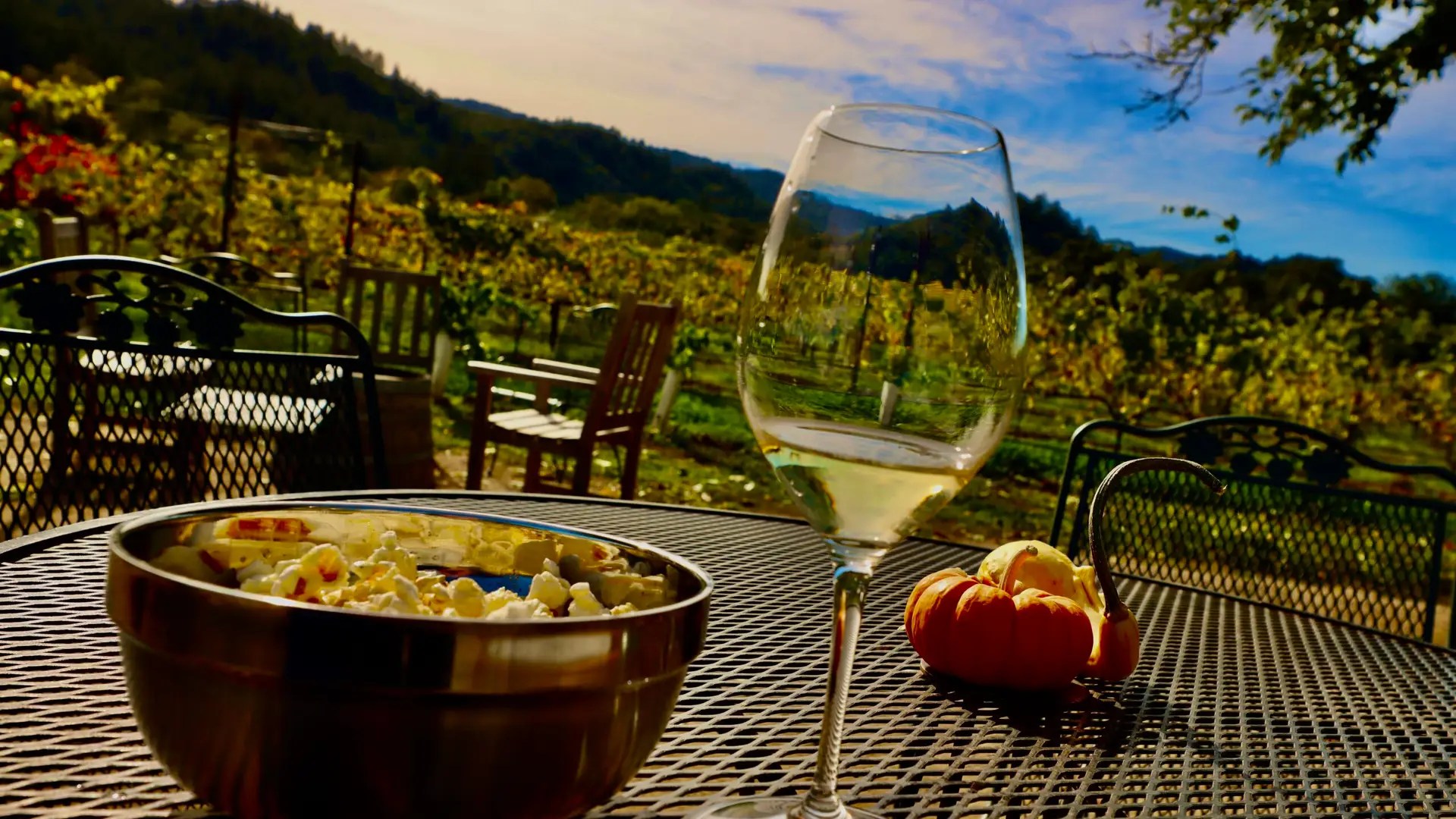 A glass of white wine and a bowl of seasoned popcorn on a patio table at Amista Vineyards, with the estate vineyard and wooded ridge behind, Dry Creek Valley, Healdsburg, Sonoma