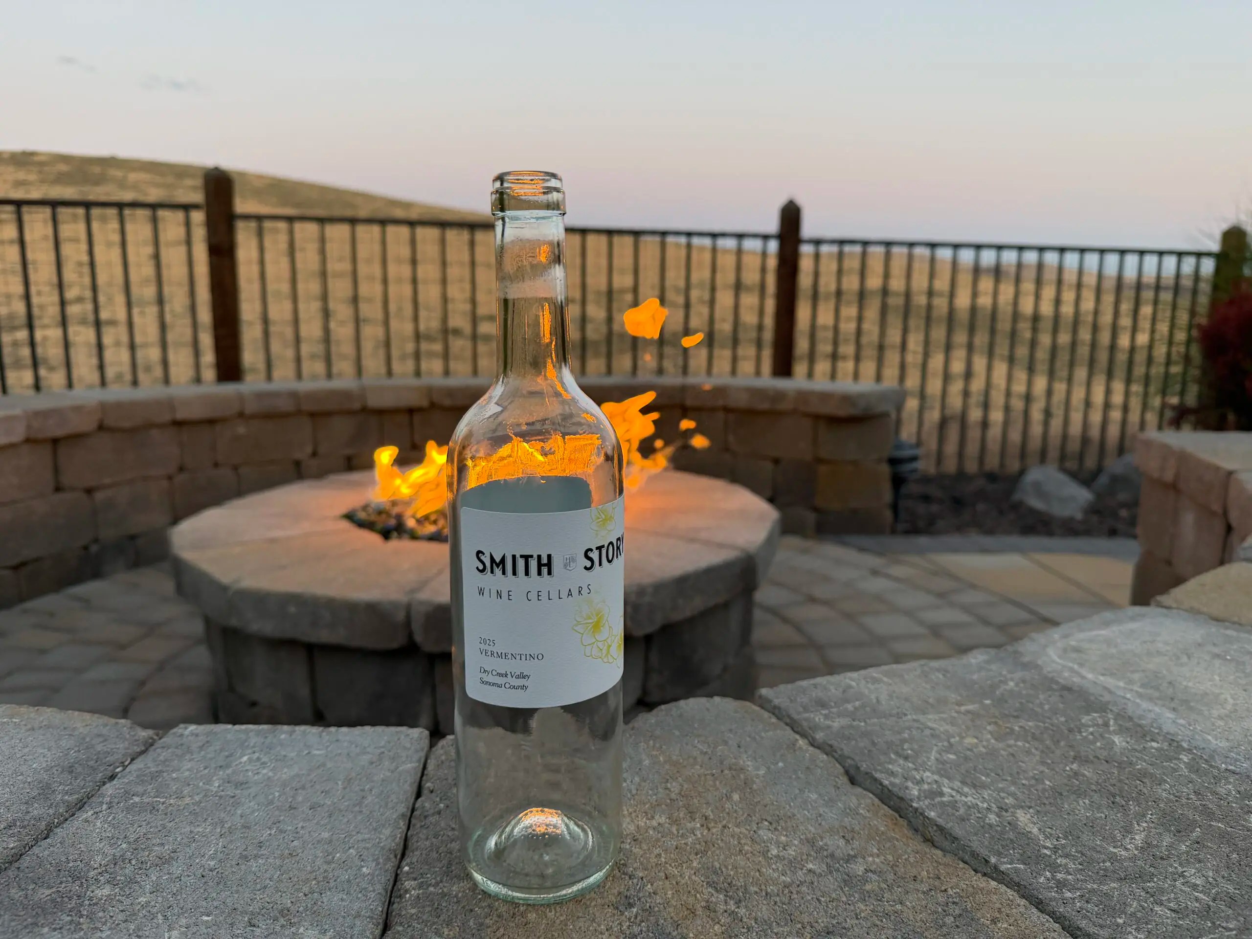 Empty bottle of Smith Story Wine Cellars 2025 Vermentino, Dry Creek Valley, standing on pavers beside a lit firepit at dusk