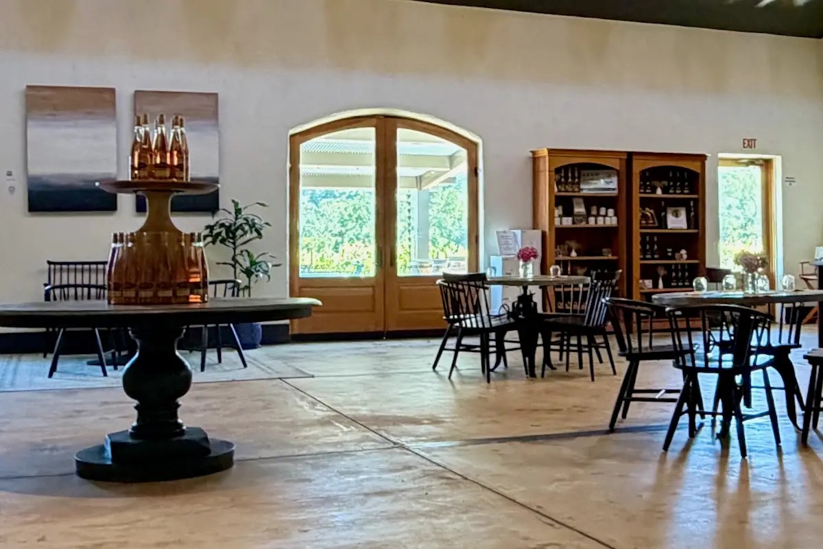 An empty winery tasting room with scattered tables and chairs, open doors looking out to vines.