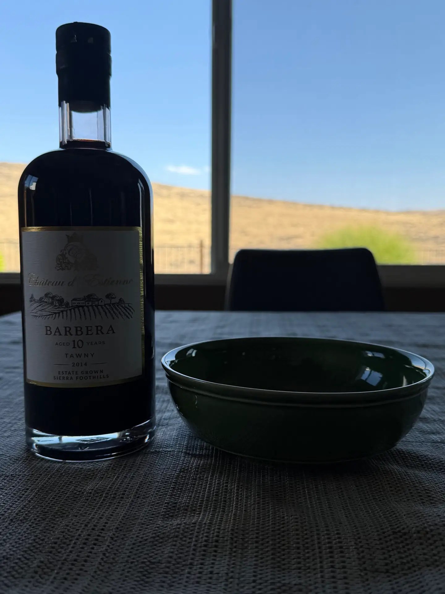 A bottle of Château d'Estienne 2014 Barbera Tawny port beside an empty dark green bowl on a table, with dry Nevada hills visible through the window behind them.