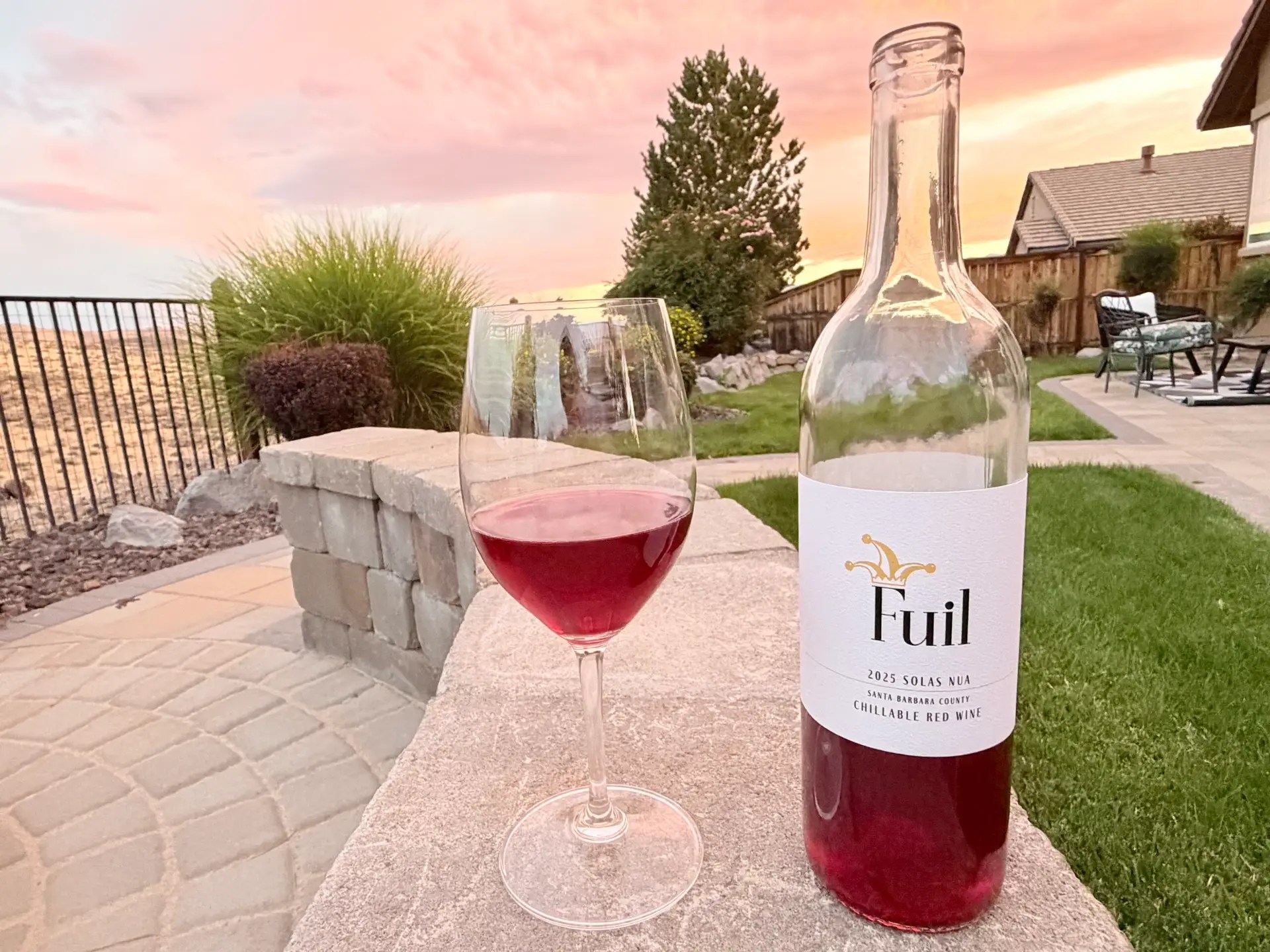 A glass of Fuil 2025 Solas Nua chillable red wine beside the bottle on a stone patio wall at sunset, with high desert hills beyond a backyard fence.