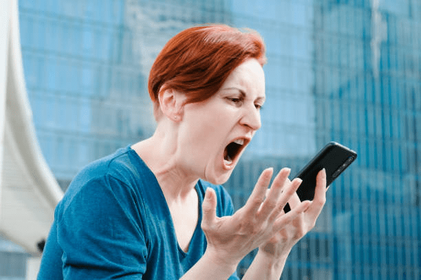 A woman with short red hair is yelling into her smartphone while expressing frustration outdoors, with a modern building in the background.