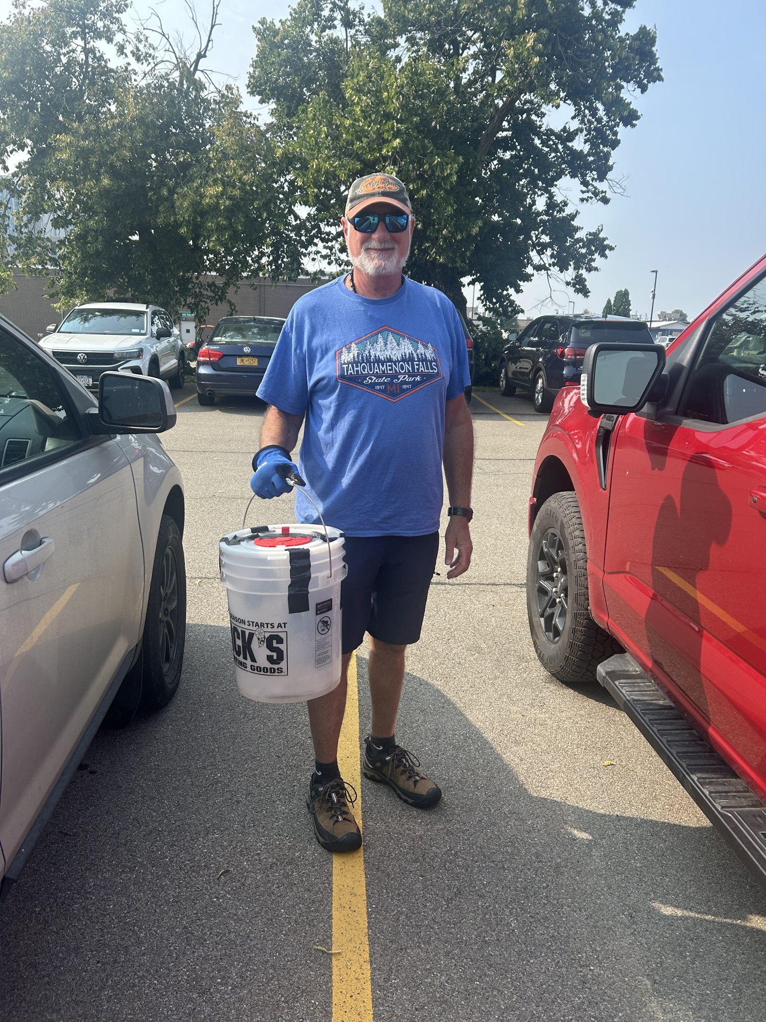 A man wearing a blue T-shirt, sunglasses, and gloves holds a bucket labeled 'Dick's Sporting Goods' while standing in a parking lot with trees in the background.
