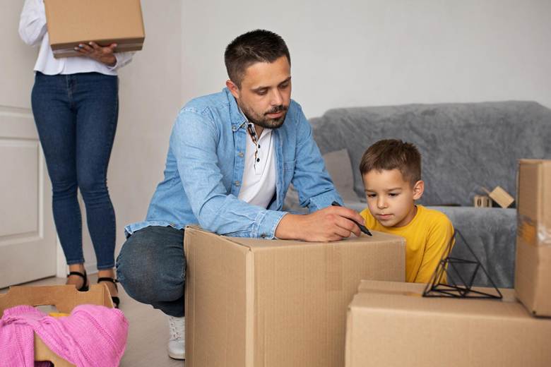 A father and his young son are sitting among moving boxes, with the father writing on a box while the son watches intently. A woman is seen in the background carrying a box.