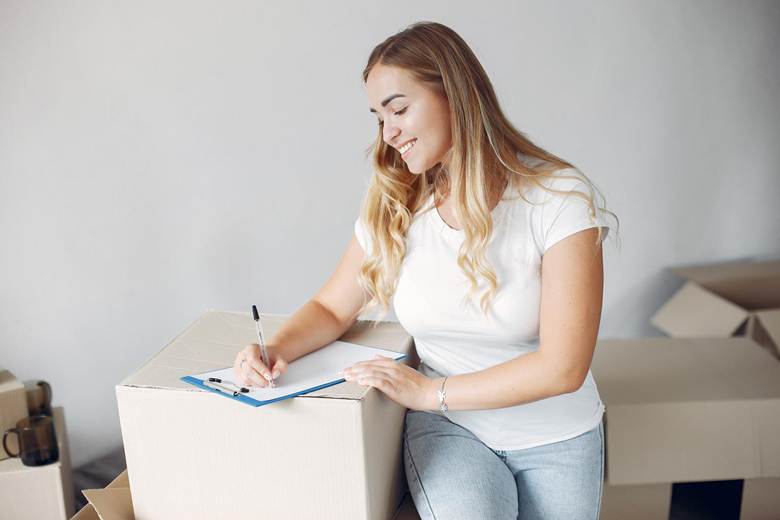 A woman with long hair is sitting on a cardboard box, smiling while writing on a clipboard with a pen.