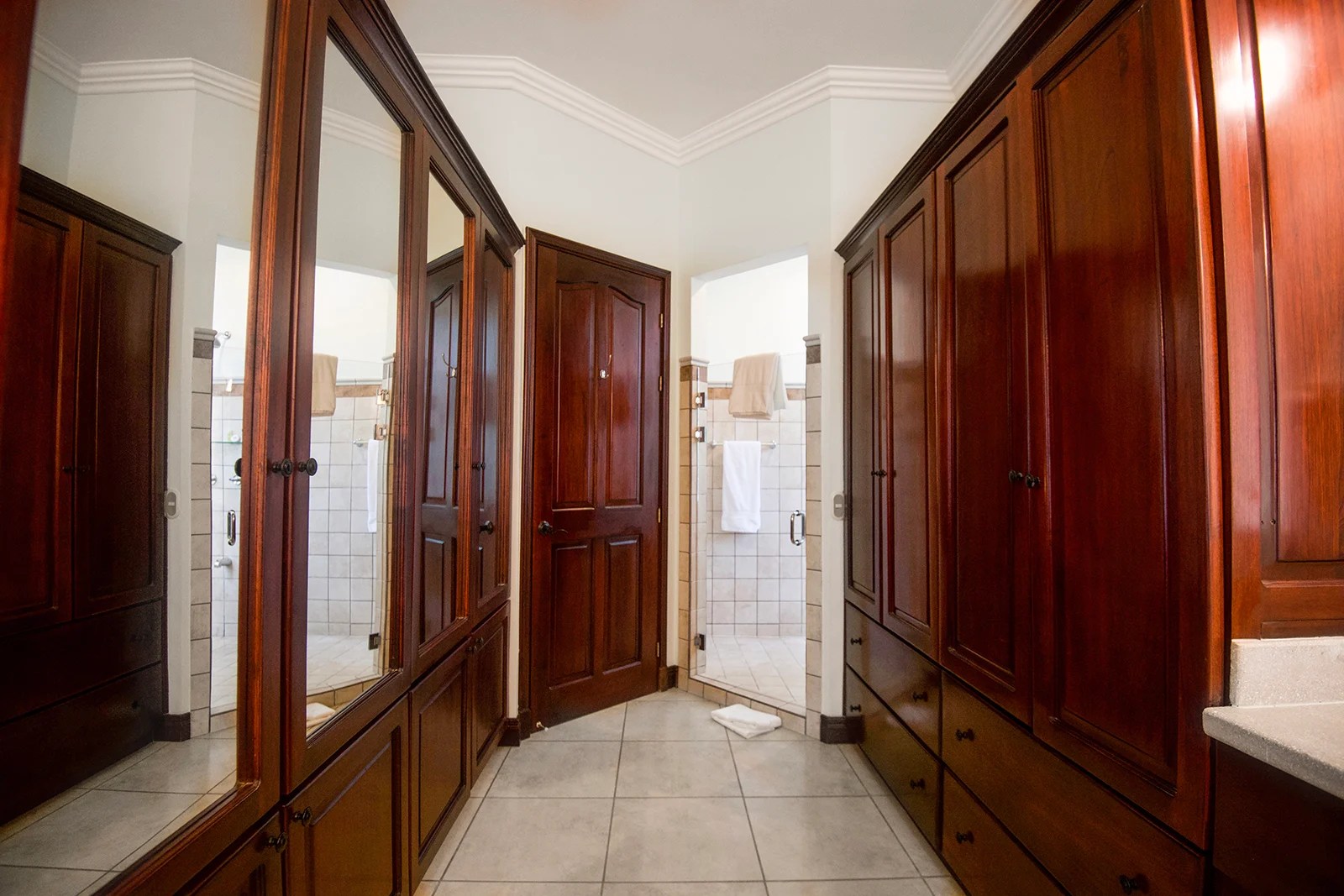 Walk-in closet with built-in wooden wardrobes and mirrors leading to an ensuite bathroom with tiled shower in a Los Sueños villa.