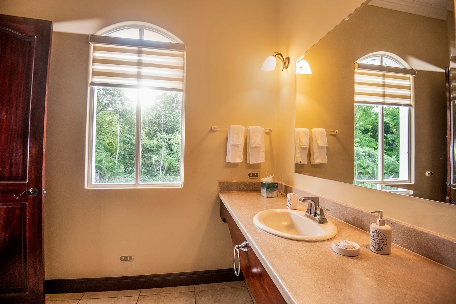 Bathroom with vanity sink, large mirror, and window looking out to jungle greenery in a Los Sueños villa.