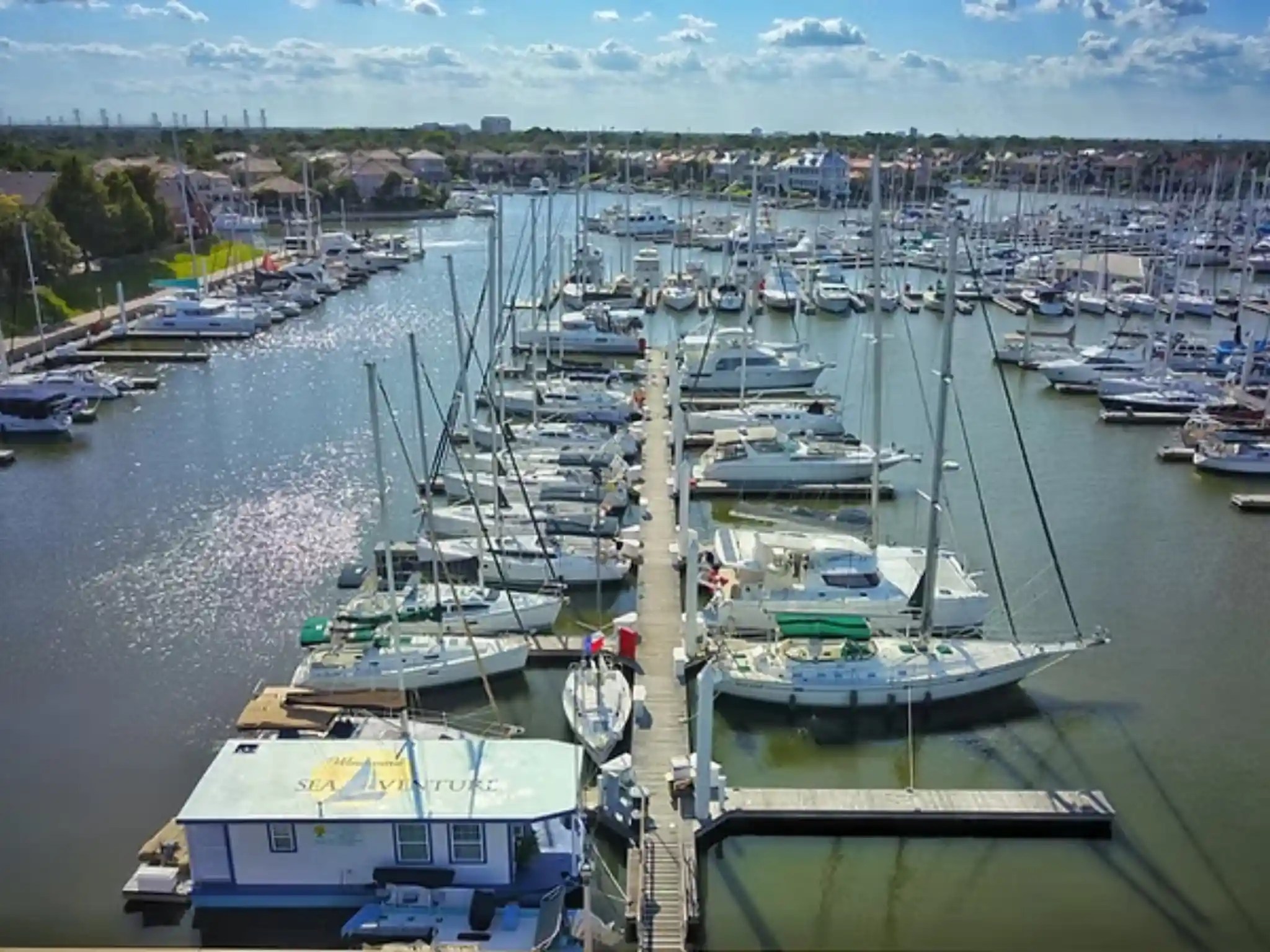 Aerial view of a marina filled with numerous sailboats docked in slips on a sunny day with blue skies and clouds above.