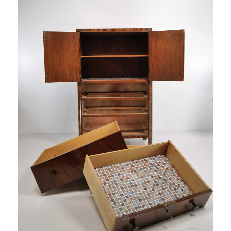 Tall wooden cabinet with two open doors revealing multiple shelves and two lower drawers; a mosaic-tiled tray rests on the floor in front of it.