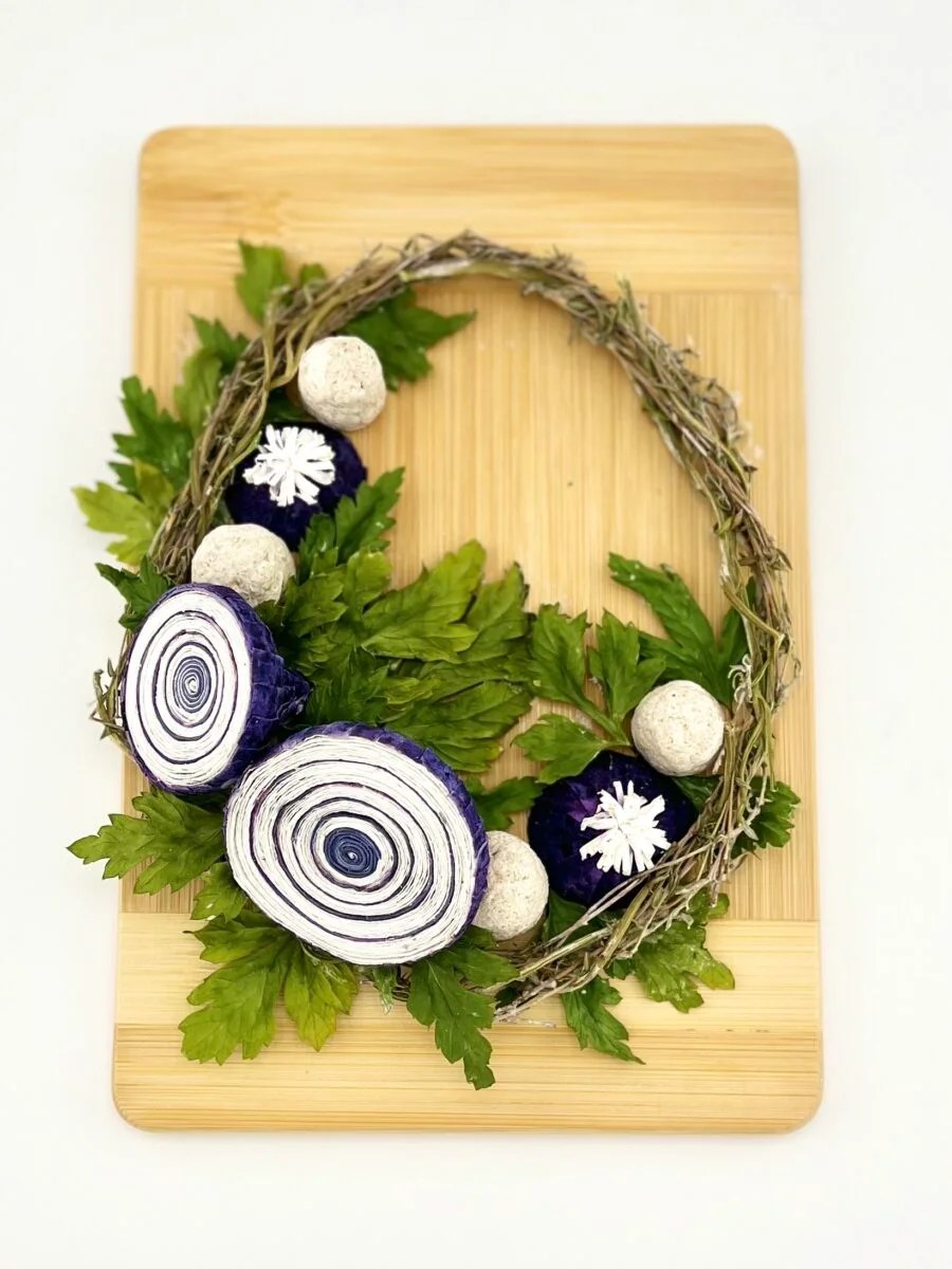 A wreath of dried herbs decorated with paper onion ends and mushroom stalks, set on a wooden cutting board.