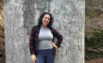 Against the backdrop of arid Mexican terrain, environment volunteer in Mexico Ilana Hirschfeld stands confidently beside a towering concrete water cistern that transformed this community's access to clean water. Her warm smile radiates pride and satisfaction as she poses with one hand on her hip, wearing a burgundy and black plaid flannel over a fitted gray top, her dark curly hair catching the natural light. The dedicated volunteer showcases the substantial cylindrical tank rising from the earth like a monument to community development, its smooth gray concrete walls creating a striking contrast with her vibrant plaid shirt. This impressive USAID-funded infrastructure project represents months of collaborative effort in sustainable water solutions, bringing hope to local families while reflecting the practical yet deeply personal nature of her environment work in rural Mexico.