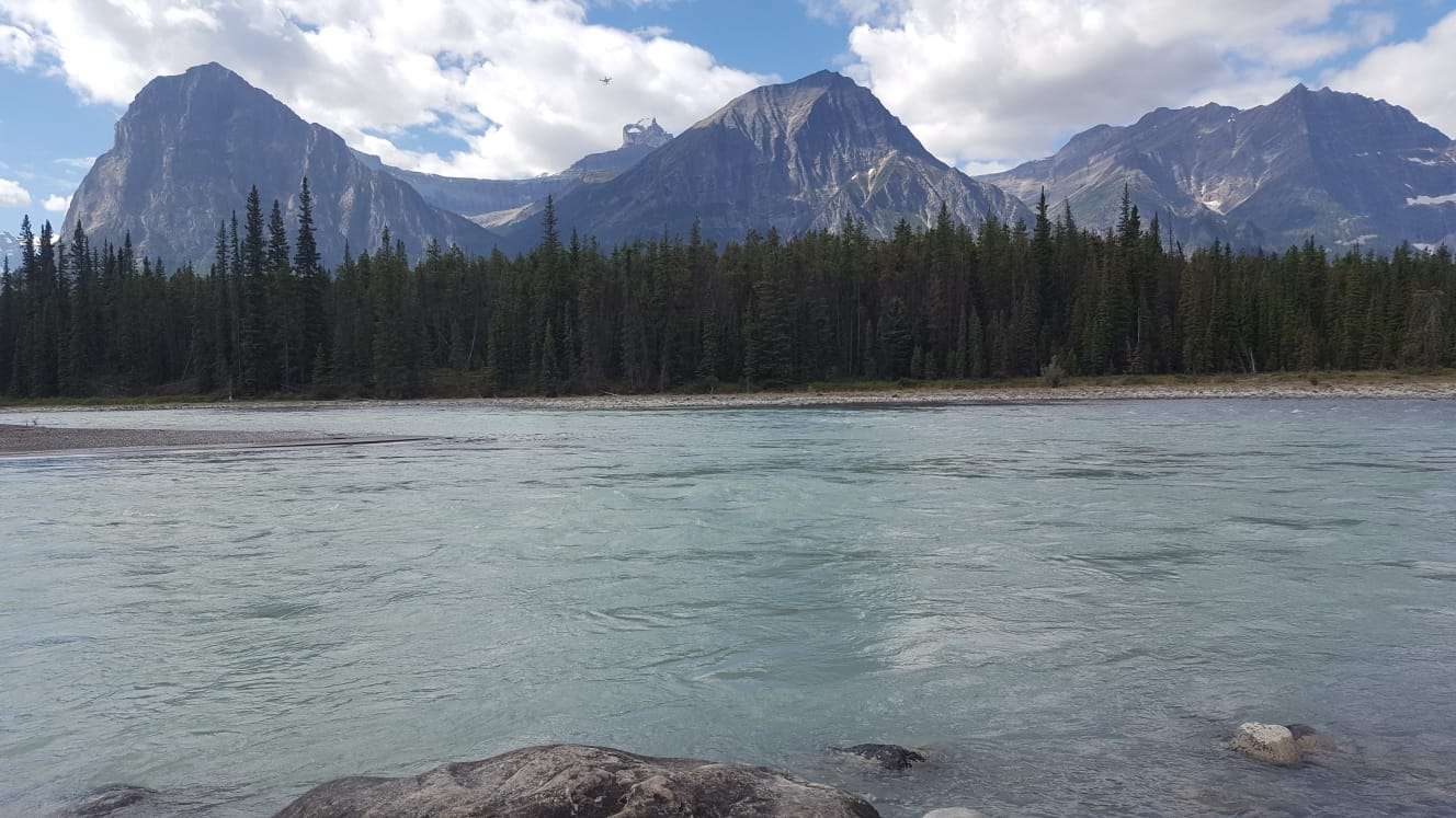 Clear blue lake overlooked by a line of evergreen trees with three mountain peaks in the background 