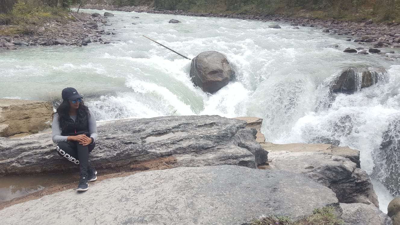 Individual wearing a black hat, black pants, and grey sweater sitting on a large grey rock with mint green water falls in the background.