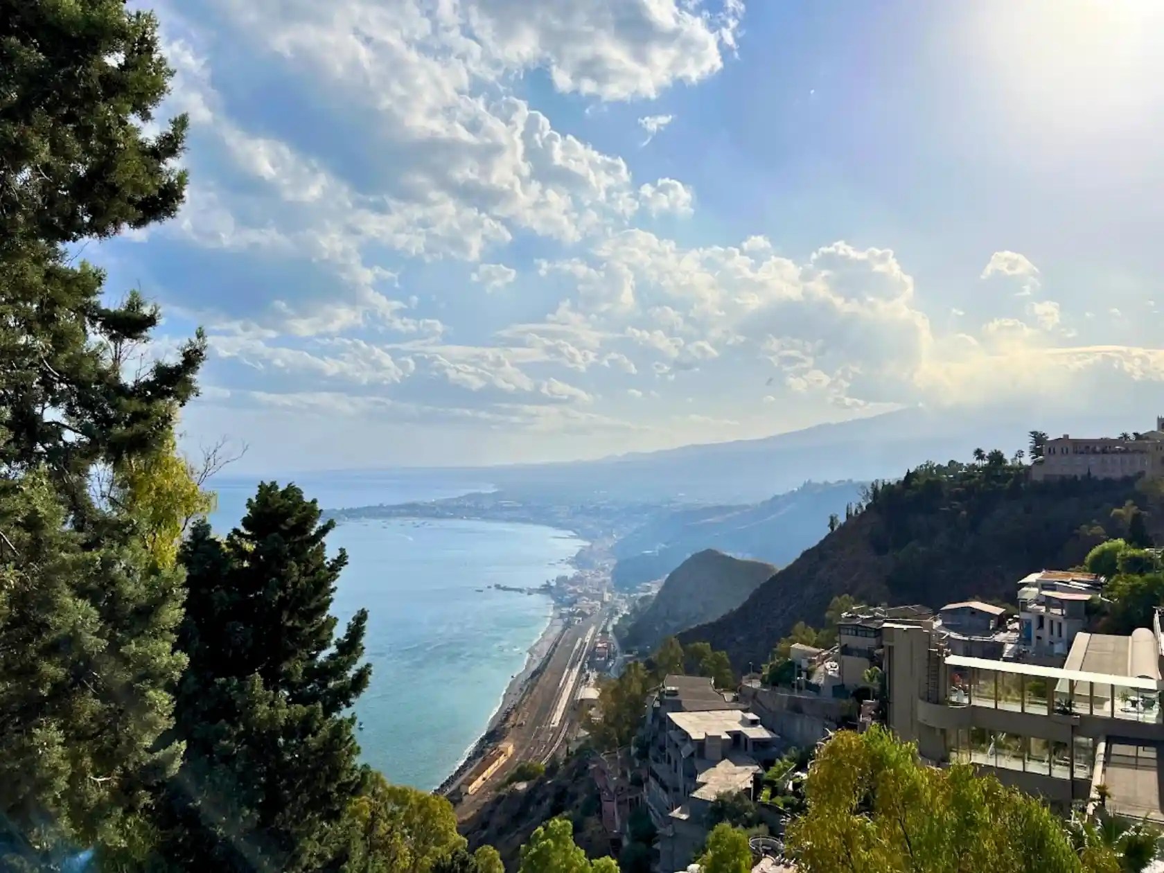 Scenic coastal view in Sicily, Italy, with the coastline stretching along the sea, framed by trees and buildings.