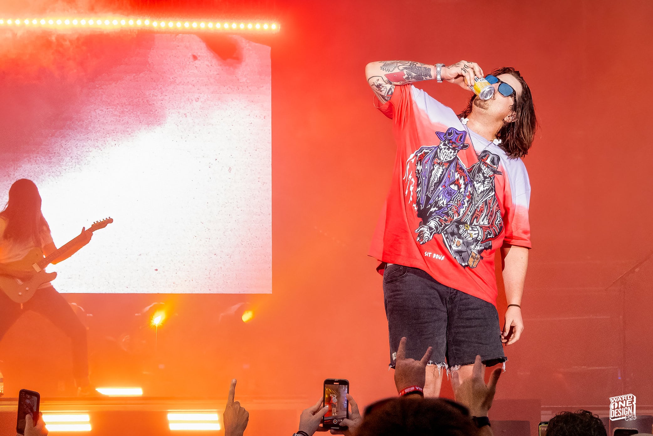Female singer with short hair and sunglasses performing on stage at Hardy Chicago, holding a microphone, with a guitarist in the background and vibrant stage lighting.
