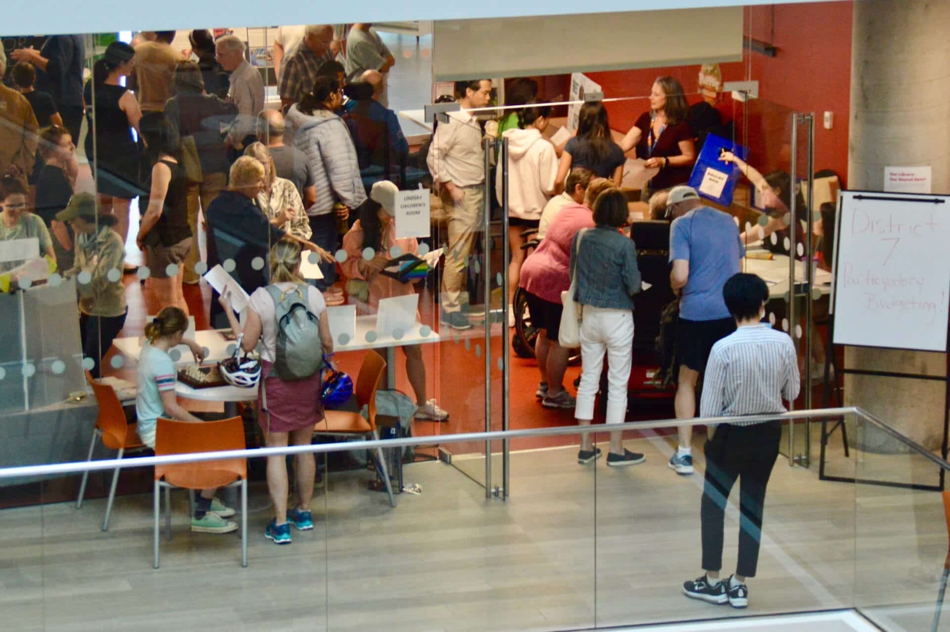 A picture of the entrance of the Lindsay Childrens room at the Central Library. To the right of the door a flip chart reads "District 7 Participatory Budgeting!" Inside through the glass walls the room is packed with people there to vote and applicants presenting their projects.
