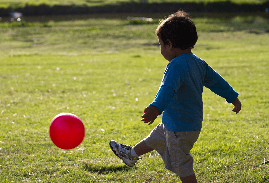A child wearing a blue long-sleeve shirt and beige shorts kicks a red ball on a grassy field. The open space and soft lighting suggest outdoor play in a park.