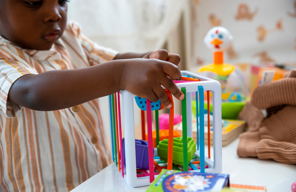 A child uses both hands to play with a colorful cube-shaped educational toy featuring rods and geometric shapes.