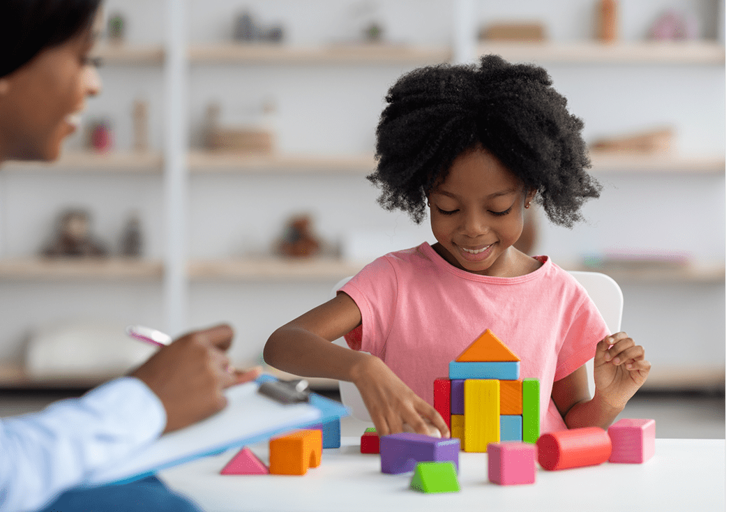 A young child builds a colorful structure with geometric blocks at a table while an adult nearby observes with a clipboard.