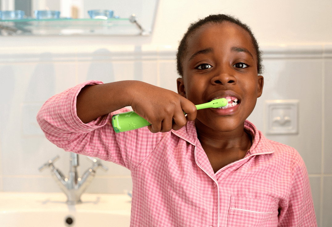 A child in pink checkered pajamas stands in a bathroom brushing their teeth with a green toothbrush. They are focused on cleaning their upper teeth.