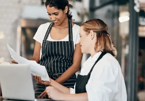 Two restaurant operators in aprons reviewing accounting documents and financial data on a laptop with TRIS