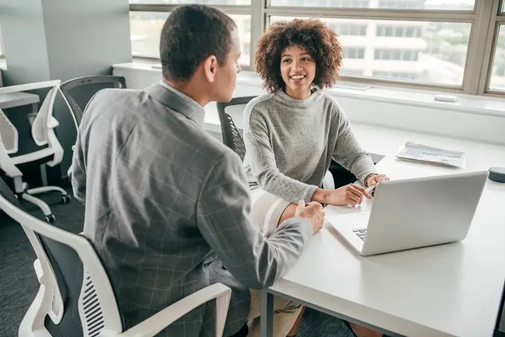 A young and smiling African American woman with curly hair sits in a chair next to her hispanic male boss having a conversation in front of her computer.