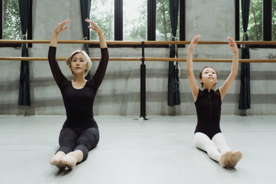 professional asian ballerina with beginner exercising in studio sitting on floor with raised arms