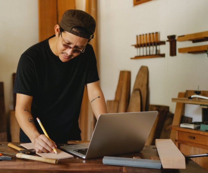 focused carpenter making notes while using laptop in studio: How to Make Money Online from Woodworking (Guided Steps)