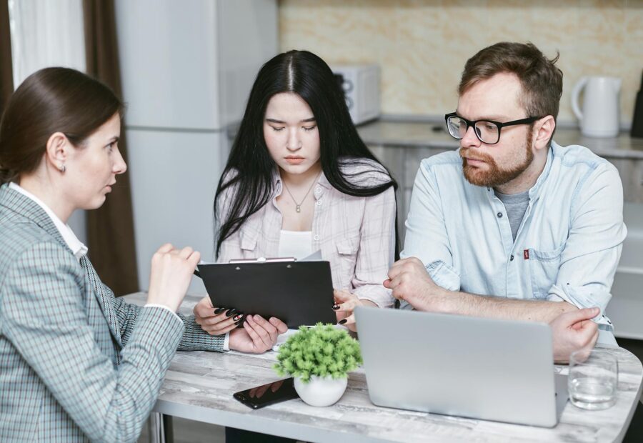 Three professionals in a meeting with one showing a clipboard to the others near a laptop.