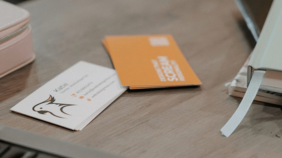 Two sets of business cards for WebDesignToo on a table next to a Macbook and notebooks