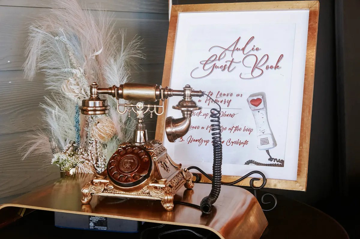 Classic Audio Guestbook A vintage-style gold telephone sits on a wooden table, accompanied by a floral arrangement of pampas grass and dried flowers. A framed sign behind reads 'Audio Guest Book' with instructions for leaving a message, featuring a graphic of a telephone handset with a heart symbol. Ideal for adding a nostalgic and interactive touch to a wedding event.
